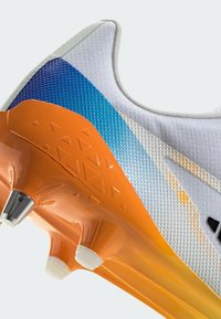 Zapatilla deportiva con una parte superior blanca texturizada con un degradado azul, suela naranja y acentos triangulares. Incluye una punta metálica en la suela.