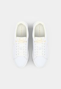 Tênis brancos feitos de material sintético liso, com atacadores finos, detalhes em logo dourado, bico arredondado e um design minimalista.
