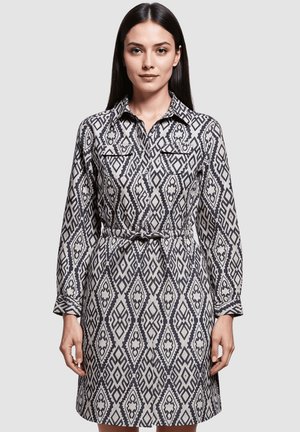 Vestido camisero con un patrón geométrico en blanco y negro, con mangas largas, un cuello, dos bolsillos en el pecho y una cintura ceñida por un cinturón.