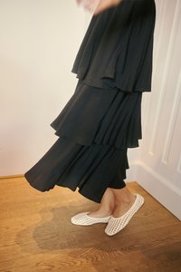Femme portant une jupe noire à étages et des chaussures plates blanches tissées, debout sur un sol en bois près d'une porte blanche.