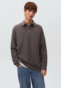 Junger Mann mit kurzen braunen Haaren, der ein lockeres graues Langarm-Polohemd über einem weißen T-Shirt und blauen Jeans trägt, mit einer Hand in der Tasche.