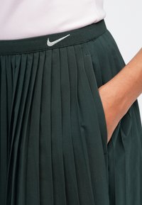 Tumši zaļa plānotā svārki ar pieguļošu jostasvietu un sānu kabatu. Uz jostas ir sudraba Nike logo, izgatavots no viegla, gluda auduma.