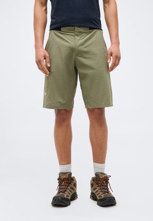 Άντρας που φοράει ελιάς πράσινα σορτς μέχρι το γόνατο, γκρι κάλτσες και καφέ παπούτσια πεζοπορίας, όρθιος μπροστά σε απλό ανοιχτό φόντο.