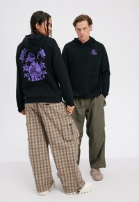 Dve osebi, oblečeni v črne puloverje s kapuco, okrašene s purpurnim napisom "Nikoli ne glej nazaj", v kombinaciji z ohlapnimi karo hlačami in olivno zelenimi cargo hlačami, stojita pred belim ozadjem.