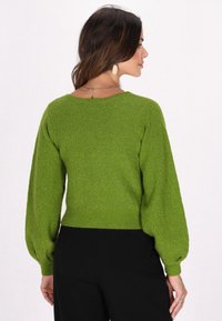 Grüner, texturierter Strickpullover mit Ballonärmeln, flachen Ausschnitt und geripptem Saum. Kombiniert mit schwarzen Hosen, die einen subtilen Schimmer aufweisen.