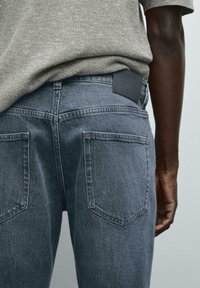 Jeans in denim grigio con vita alta, due tasche posteriori e un'etichetta in pelle nera. Il tessuto ha una finitura leggermente strutturata.
