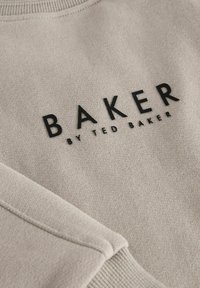 Sweatshirt beige clair avec un col rond, des poignets côtelés et un logo noir brodé indiquant "BAKER by TED BAKER". Texture lisse.