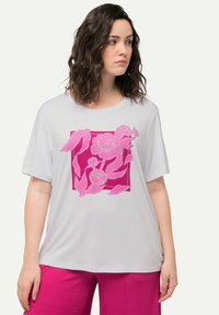 Camiseta gris claro con un gran estampado floral en rosa sobre un fondo cuadrado. Mangas cortas y un corte relajado. La tela parece suave.