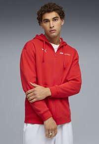 Rød zip-up hættejakke lavet af blød stof, med lange ærmer, elastiske manchetter, og en justerbar hætte. Puma-logo og "Formula 1" branding visuelt præsenteret.
