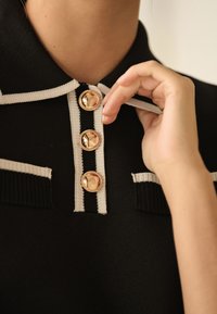 Schwarzes Polohemd mit weißen Akzenten, ausgestattet mit drei großen goldfarbenen Knöpfen und einer gerippten Textur. Eine Hand berührt den Kragenbereich und betont das Design.