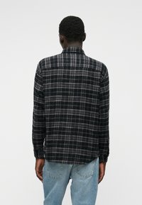 Μαύρο και γκρι plaid φανέλα, μακριά μανίκια, γιακάς με κουμπιά, ελαφρώς κυρτή στρίφωση, φαίνεται από πίσω. Φορεμένο με ανοιχτό γαλάζιο τζιν.