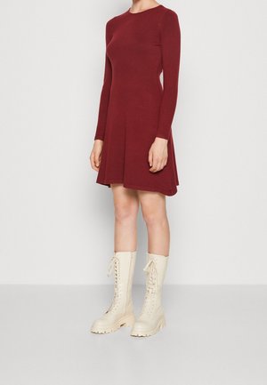 Woman wearing a long-sleeve burgundy dress paired with knee-high cream lace-up boots, standing against a plain light background.