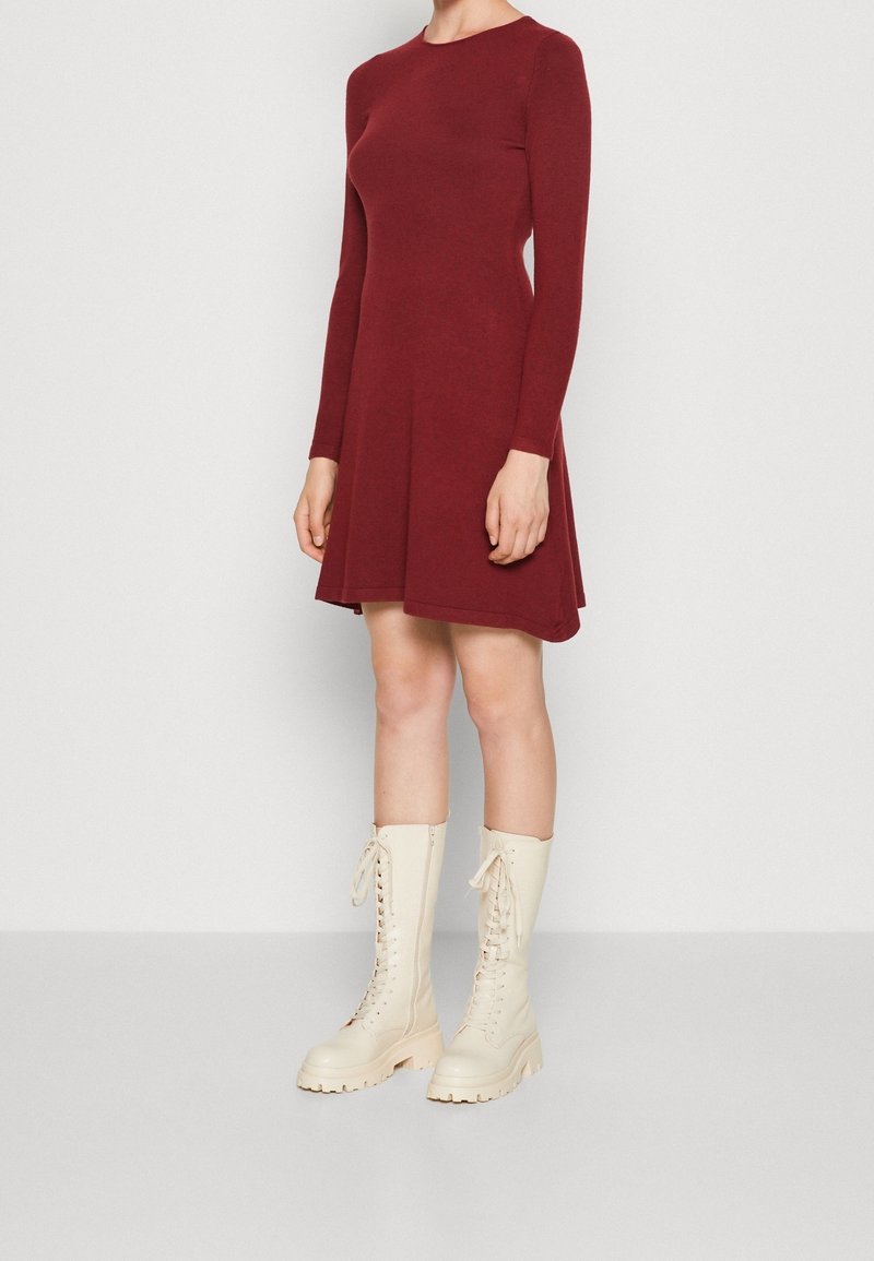Femme portant une robe bordeaux à manches longues associée à des bottes lacées crème montant jusqu'au genou, debout devant un fond clair uni.