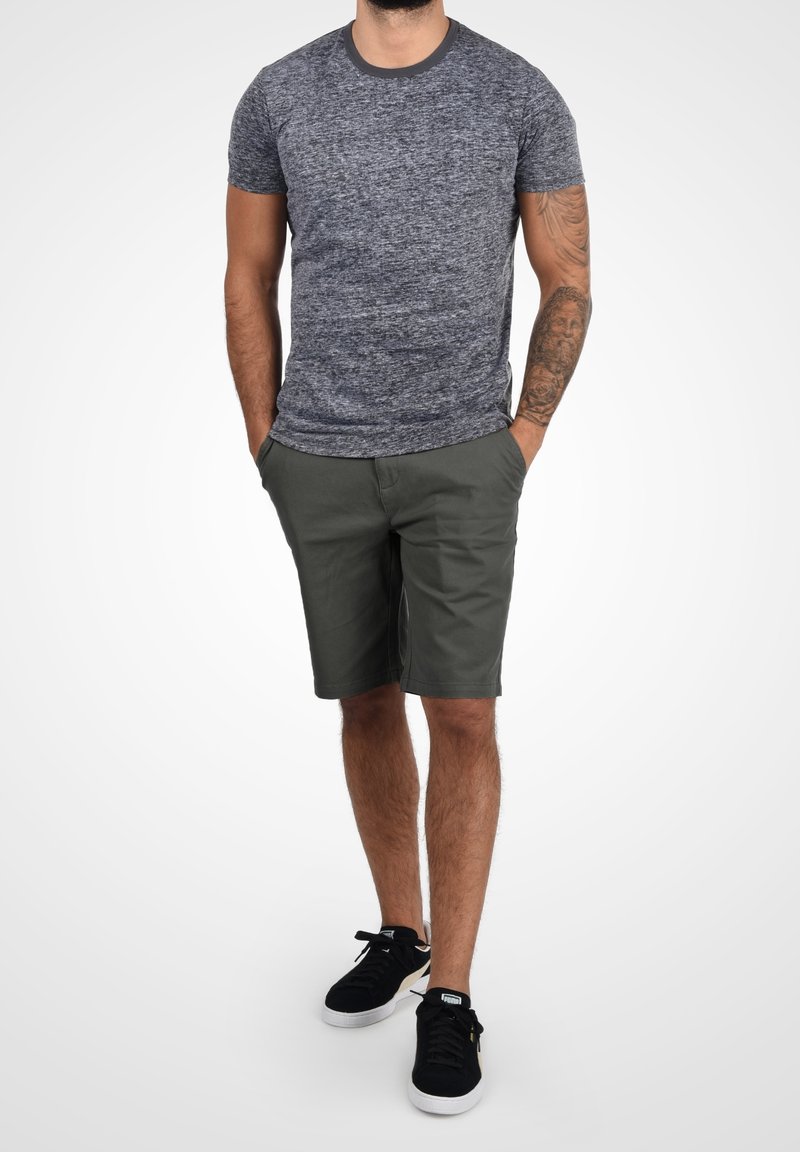 Homme debout les mains dans les poches, portant un t-shirt gris chiné, un short vert olive, des baskets noires, et un tatouage sur l'avant-bras gauche.