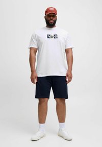 Hvid bomulds t-shirt med grafisk motiv på forsiden, kombineret med marineblå shorts og hvide sneakers. Modellen har en rød kasket på. Enkel, afslappet outfit.