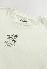 Camiseta de algodón color crema con un diseño floral negro y el texto "Jardines de Serenidad Silvánica" debajo. Cuello redondeado y mangas cortas.