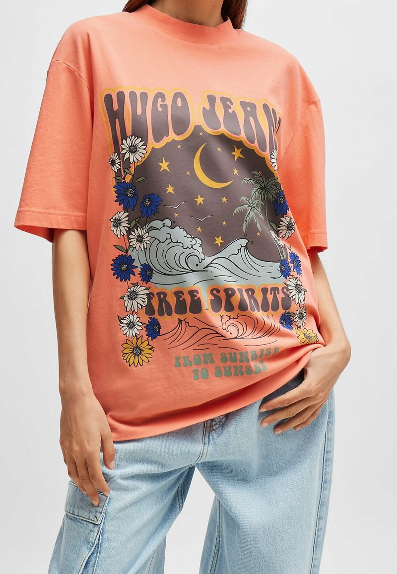 Camiseta de gran tamaño color coral hecha de algodón, con un estampado gráfico de olas, flores y el texto "ESPÍRITUS LIBRES" en letras en negrita.
