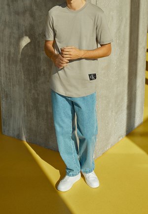 Mann trägt beige T-Shirt mit Calvin Klein-Logo, hellblaue Jeans und weiße Sneakers, steht vor einer Betonwand auf gelbem Boden.