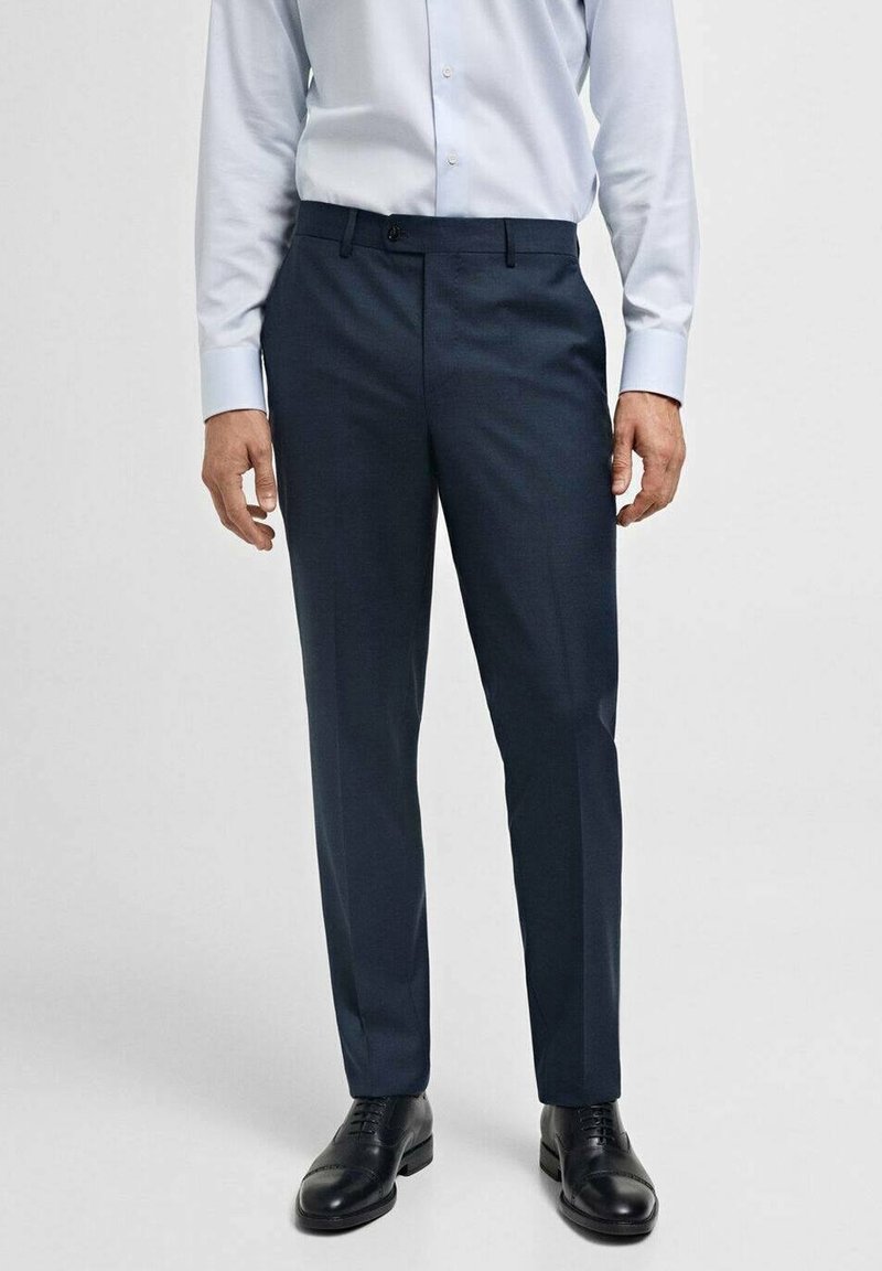 Homme portant un pantalon de costume bleu marine foncé, des chaussures en cuir noires et une chemise bleue claire à manches longues, sur un fond uni.