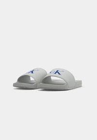 Szürke, nyitott orrú csúszóslip-on papucsok, széles pántokkal, kék "cK" logóval, és texturált talppal a tapadásért.