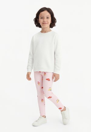 Enfant aux cheveux foncés portant un sweat-shirt blanc, des leggings roses avec des motifs de fruits et de cupcakes, et des baskets blanches, debout sur un fond blanc.