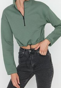 Grüner, kurz geschnittener Sweatshirt mit Reißverschluss, langen Ärmeln, gerippter Struktur und Kordelzug am Saum, kombiniert mit hoch taillierten schwarzen Jeans.