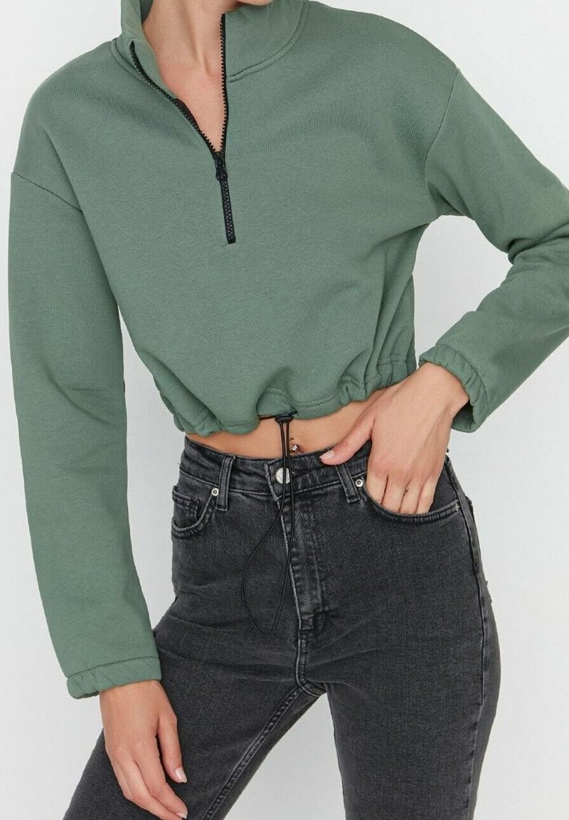 Grüner, kurz geschnittener Sweatshirt mit Reißverschluss, langen Ärmeln, gerippter Struktur und Kordelzug am Saum, kombiniert mit hoch taillierten schwarzen Jeans.