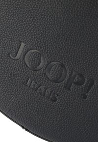 Čierna textúrovaná koža s embossovaným logom "JOOP! JEANS". Kruhový tvar, ktorý zdôrazňuje hladký povrch s jemnými vzorovými detailmi.