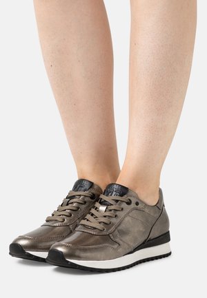 Pieds portant des baskets lacées gris métallisé avec des semelles noires et blanches, présentés sur un fond clair uni.