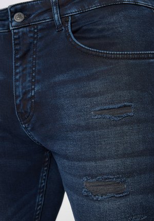 Ciemnoniebieskie dżinsy o slim fit, z przetarciami i postrzępionymi łatami. Tkanina ma gładką teksturę z delikatnym połyskiem.