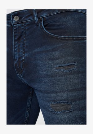 Ciemnoniebieskie dżinsy o slim fit, z przetarciami i postrzępionymi łatami. Tkanina ma gładką teksturę z delikatnym połyskiem.