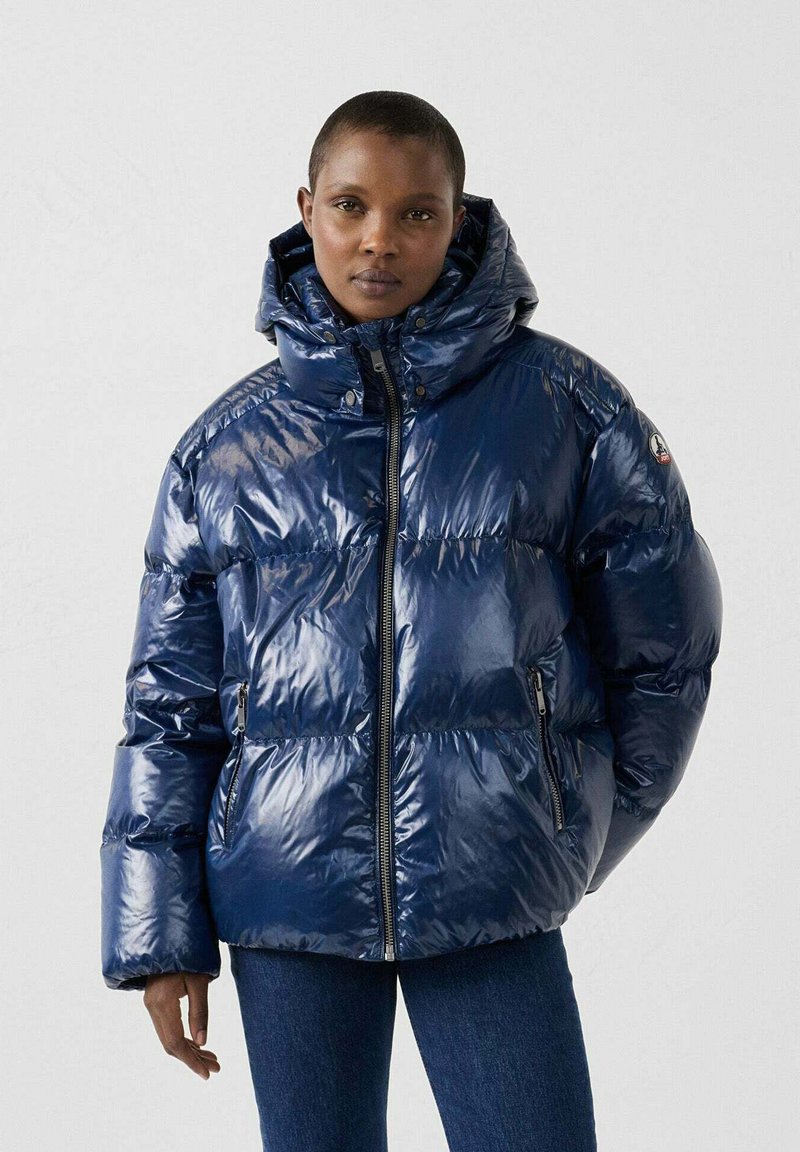 Giacca puffer blu navy con finitura lucida, cappuccio oversize, zipper frontale e tasche laterali. Presenta un motivo trapuntato e polsini a coste.