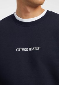 Kék árnyalatú pulóver, kerek nyakkal, fehér "GUESS JEANS" felirattal írásos betűtípussal; puha anyag, sima textúrával.