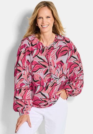 Femme souriante portant un chemisier ample à motifs roses et violets avec des manches ballon et un pantalon blanc, debout devant un fond clair.