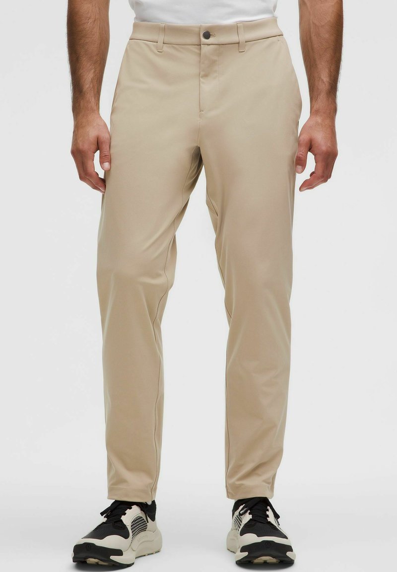 Beige színű, egyenes szabású nadrág, sima, könnyű anyagból készült. Gombos záródással és oldalsó zsebekkel rendelkezik. Fekete-fehér sportcipőkkel viselve.