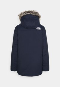 Tmavě modrá izolovaná parka s kapucí lemovanou kožešinou a logem na horní části zad. Hladká textura, dlouhé rukávy a rovný střih.