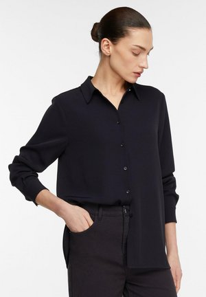 Frau mit dunklem Haar zu einem Dutt gebunden, die ein schwarzes, langärmeliges Hemd mit Knöpfen und dunkle Hosen trägt, mit einer Hand in der Tasche, schaut seitlich nach unten.