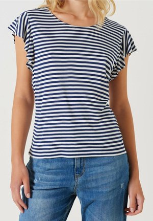 Kobieta ubrana w granatowo-biały krótki top w paski z falbanami na rękawach, do tego niebieskie dżinsy.