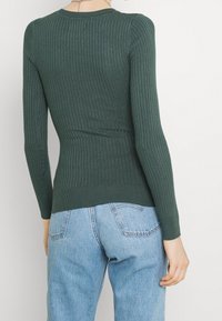 Ribbed pitkähihaine villapaita tummanvihreänä, jossa on pyöreä kaula-aukko ja istuva muotoiluelementti. Yhdistetty vaaleansinihin farkkuihin.