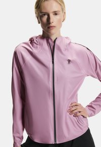 Veste de sport rose clair avec une fermeture éclair complète, une capuche et des accents noirs contrastants sur les épaules. Tissu lisse et léger.