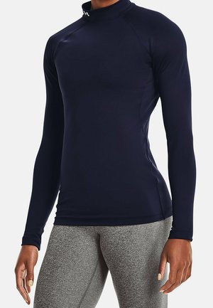 Frau trägt ein eng anliegendes, langärmliges, marineblaues Sportoberteil und graue, strukturierte Leggings, steht mit locker an den Seiten hängenden Armen.