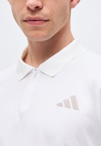 Jeune homme portant un polo blanc avec un petit logo Adidas clair sur la poitrine gauche et un col partiellement zippé.