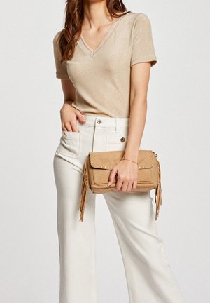 Vrouw die een beige top met korte mouwen en een witte broek draagt en een beige gestructureerde clutch met tassels en studs vasthoudt.
