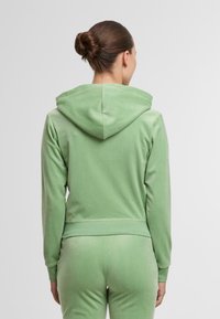 Zöld velúr kapucnis felső, testhezálló kialakítással, húzózsinóros kapucnival és hosszú ujjal, egy modellen, hátulról bemutatva.