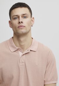 Lys rosa polo-skjorte med teksturert vev, to knapper på plagget, og en liten logo på brystet. Korte ermer og krage-design.