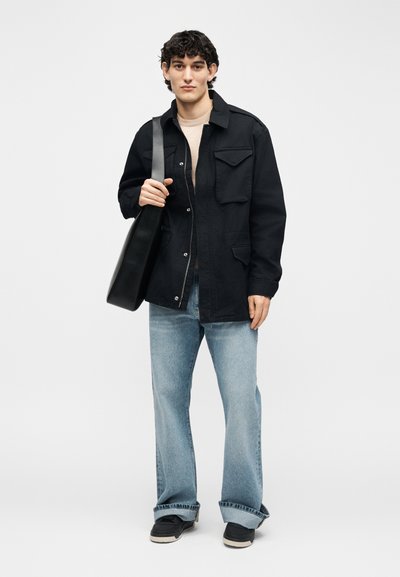 Νεαρός άνδρας φορώντας μαύρο μπουφάν, μπεζ πουκάμισο, ανοιχτό μπλε τζιν, μαύρα αθλητικά παπούτσια, κρατώντας μια μαύρη τσάντα ώμου, στέκεται μπροστά σε απλό φόντο.