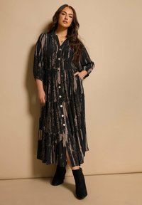 Lang schwarzes Kleid mit beigen abstrakten Streifen, durchgehender Knopfleiste, gestuftem Design und Taschen; kombiniert mit schwarzen Knöchelstiefeln.