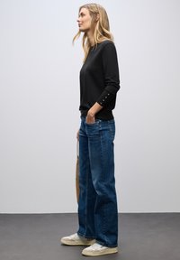 Maglione nero con maniche a tre quarti e dettagli in bottone, abbinato a jeans blu larghi e sneakers chiare. Texture liscia.