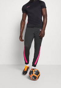 Sort Nike t-shirt med korte ærmer og pink detaljer, parret med grå sweatpants med pink sidelister. En sort og orange fodbold ved fødderne.
