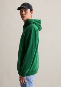 Jonge man in een groene hoodie, lichtblauwe jeans en een zwarte pet, zijwaarts staand tegen een effen lichte achtergrond.
